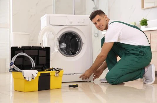 Technician in green overalls inspecting a front-loading washing machine beside a yellow toolbox